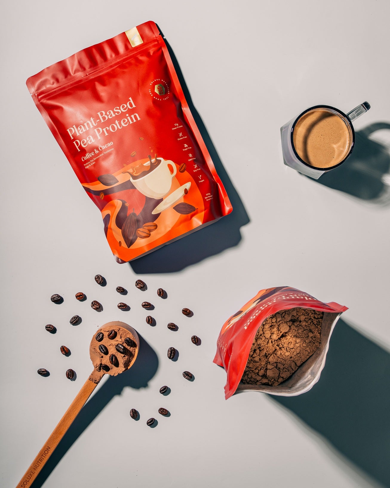 Red protein powder package in chocolate protein powder series with coffee beans, a wooden spoon, and a cup of coffee on a light gray background.